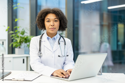 Portrait of a serious young African American female doctor in uniform sitting at a desk, working on a laptop and looking confidently at the camera