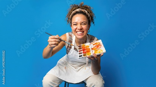 Young woman artist holding paintbrush and watercolor palette on blue background