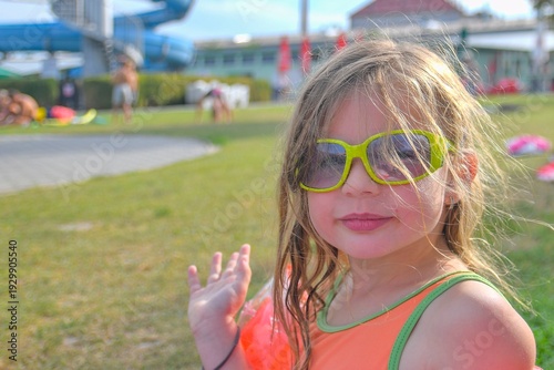 Young girl with sunglasses waving hello at a water park on a sunny day. A happy child wearing bright sunglasses and an orange swimsuit enjoys a summer day at a busy water park with slides in the