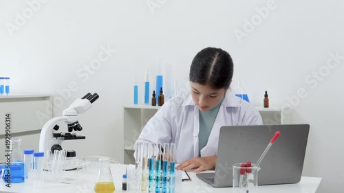 Young Asian women Scientist wearing lab coat writing a report with blue liquid during experiment in modern laboratory with microscope and test tubes rack and laptop