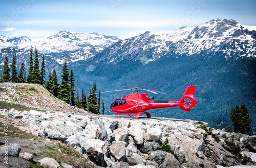 A vibrant red helicopter rests on a rocky outcrop against a backdrop of snow-capped mountains. Conifer trees and blue skies enhance the scenic view.