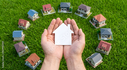Hands holding small paper house and model houses on grass