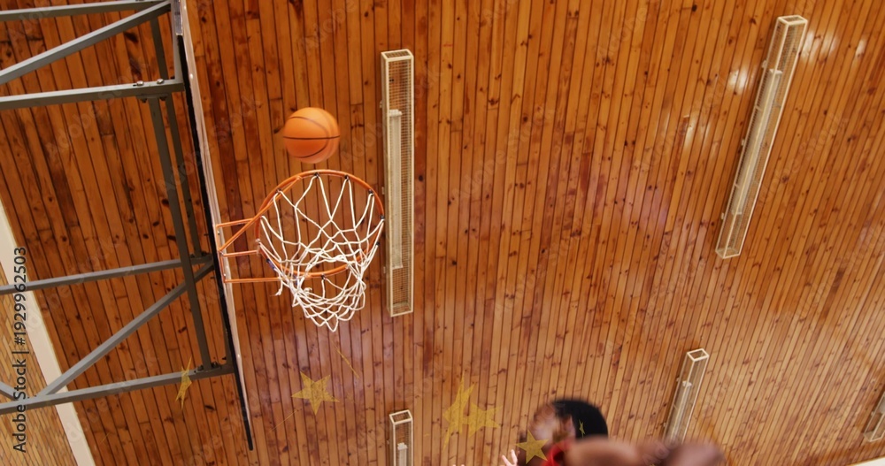 Fototapeta premium Sinking orange basketball falling through rim in school gymnasium with players' jerseys and hands