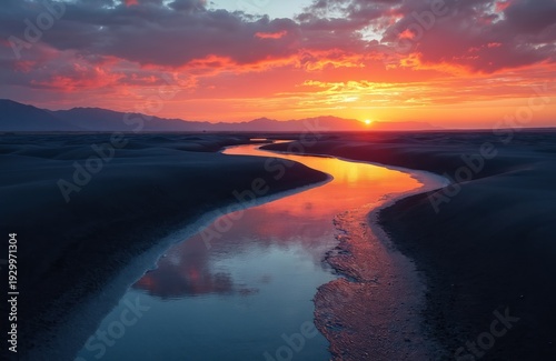 Meandering river flows through dark landscape at sunset. Water reflects vibrant orange pink clouds and sky. Distant mountains fade into horizon line.