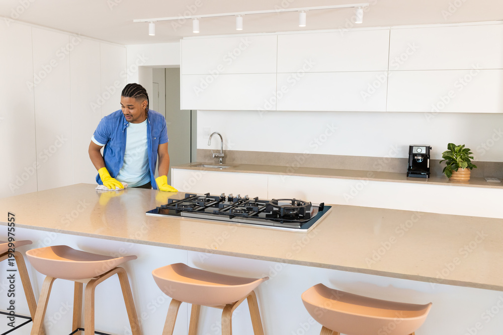 Obraz premium Man cleaning kitchen island with gas cooktop using cloth and yellow rubber gloves, copy space