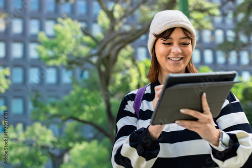 Slika na platnu Mid-adult woman in beanie tapping tablet in folio at park with stylus purple str