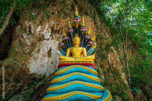 Golden Buddha with Naga statue at Wat Prangkasi in Thong Pha Phum, Kanchanaburi, Thailand surrounded by lush forest cliff