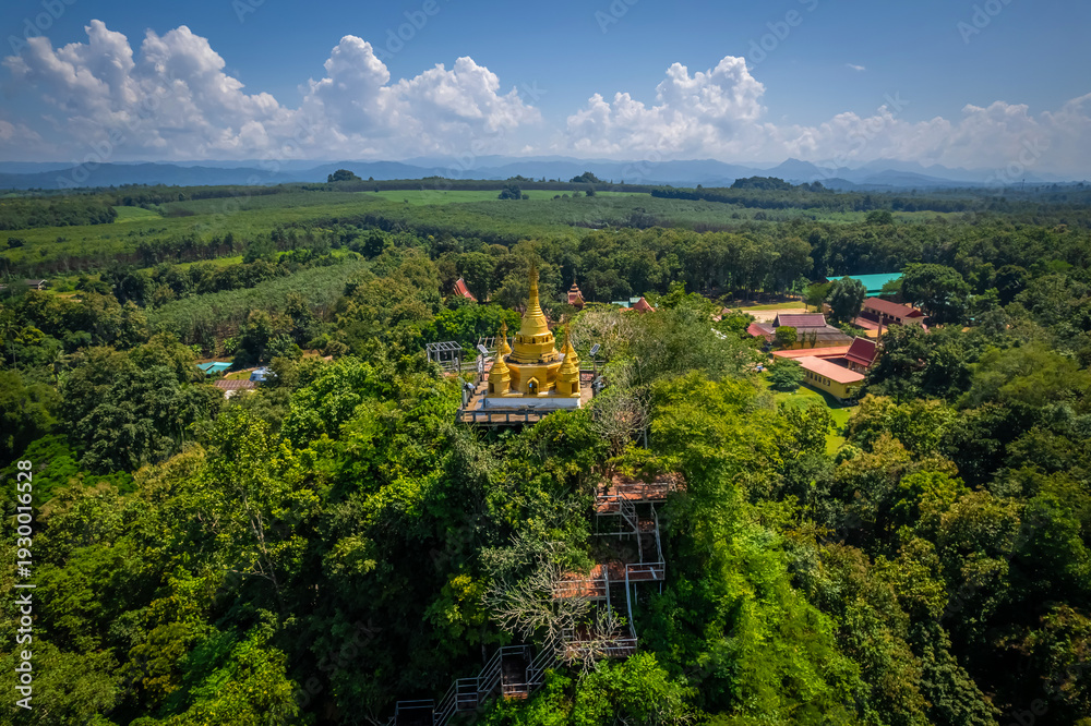 Fototapeta premium Aerial drone view of golden pagoda at Wat Prangkasi in Thong Pha Phum, Kanchanaburi, Thailand surrounded by lush forest mountain landscape