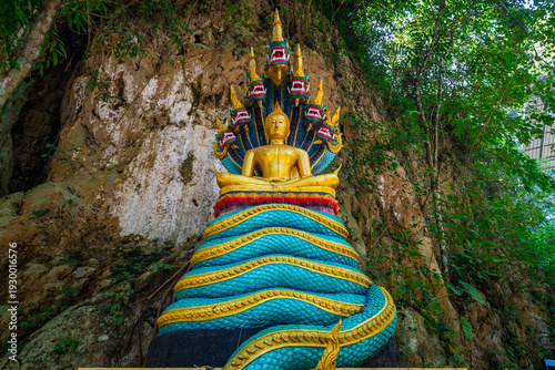 Golden Buddha with Naga statue at Wat Prangkasi in Thong Pha Phum, Kanchanaburi, Thailand surrounded by lush forest cliff