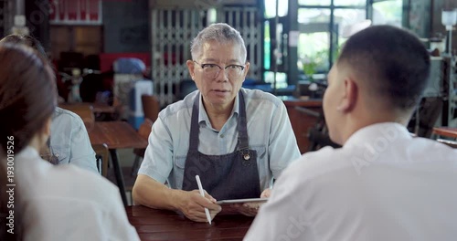 Asian senior cafe owner shares ideas with young employees, teamwork collaboration between generations creates trust, improves workflow, and supports family small business success in friendly workspace