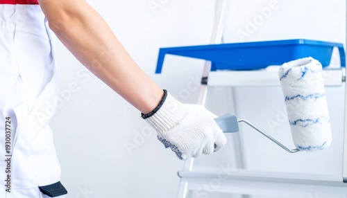 Un peintre professionnel portant des gants blancs tient un rouleau de peinture près d'un escabeau et d'un bac bleu lors de la rénovation d'un intérieur.