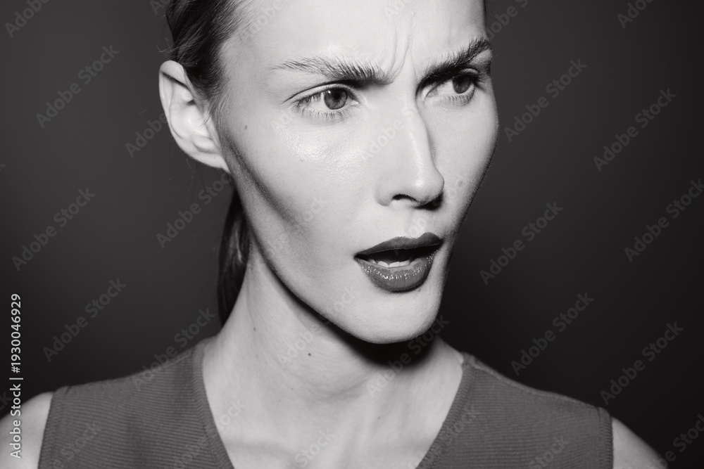 Fototapeta premium White woman with pale skin shows confused expression in black and white close-up portrait with dark background. Concept for emotion and doubt.