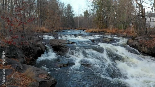 Drone flying at low altitude over the rivers with rapids staying still for a bit than moving forward over the small waterfalls with colorful trees in the Fall