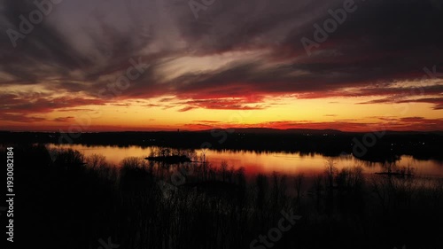 Drone crane shot moving down slowly behind the black silhouette of bare trees, at the end of sunset, with dramatic colors of orange, gold, and turquoise 