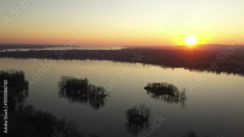 Drone shot, gently moving the camera up over calm water, dotted with small islands, showing a relaxing landscape during a sunset in a cloudless sky, behind mountains