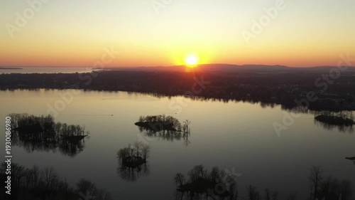 Drone shot, gently receding over calm water, dotted with small islands, showing a relaxing landscape during a sunset in a cloudless sky, behind mountains