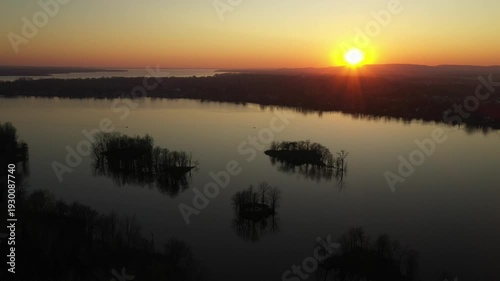 Drone shot, moving slowly over calm water, dotted with small islands, showing a sunset in a cloudless sky, behind mountains