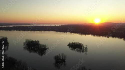 Drone shot, panning gently over calm water, dotted with small islands, showing a relaxing landscape during a sunset in a cloudless sky, behind mountains