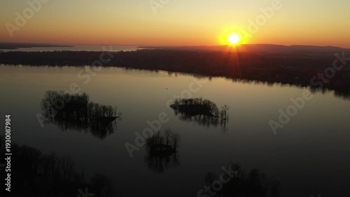 Drone shot, remaining almost stationary over calm water, dotted with small islands, showing a sunset in a cloudless sky, behind mountains