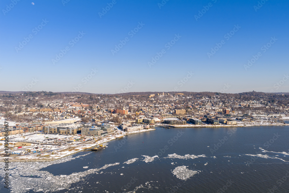 Obraz premium Winter aerial view showing urban development along a frozen riverfront, featuring many buildings, snow dusted hills, and scattered ice chunks floating on the water under a clear blue sky