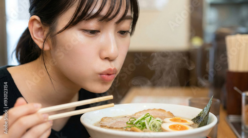 黒髪の女性が熱いラーメンを冷ます仕草・立ち上る湯気とシズル感のある一杯