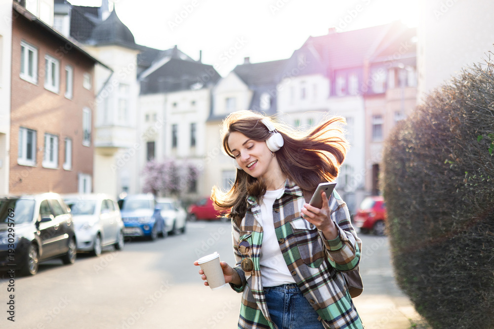 Fototapeta premium Happy woman in headphones with smartphone dancing on town street. Girl listening to music playlist outdoors, walking in city, having fun. Urban student lifestyle. Whimsical, zest for life