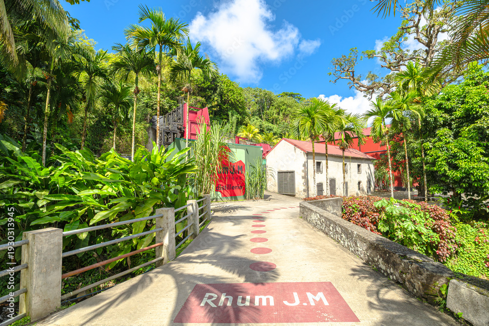Fototapeta premium Martinique, French Antilles - Dec 27, 2025: Rhum JM distillery buildings nestled in lush tropical vegetation with palm trees and a small stream reflecting a bright blue sky