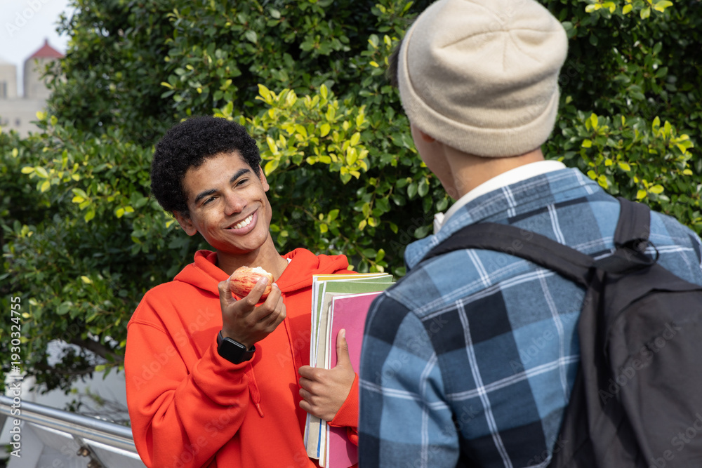 Fototapeta premium Diverse teenage male friends chatting on campus walkway, orange hoodie holding apple and notebooks