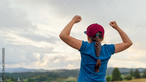 Rear view of a girl in a cap raising her arms in a gesture of victory and empowerment, standing outdoors against a scenic landscape and cloudy sky. Concept of success, confidence, and achievement.