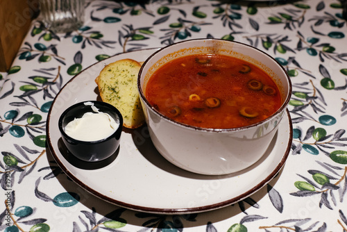 Bowl of traditional Russian Solyanka soup with olives, served with garlic bread and sour cream. Rich and spicy meat soup on a floral tablecloth in a restaurant setting.