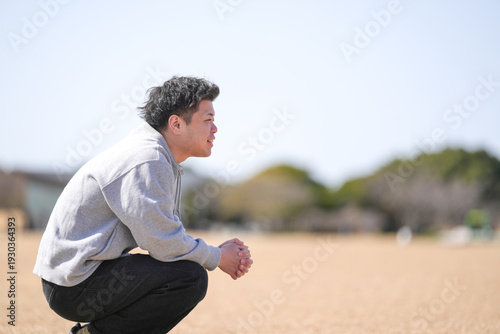 A man is kneeling down in a field and looking up at the sky. He is wearing a gray hoodie and black pants. Scene is peaceful and contemplative, as the man seems to be lost in thought