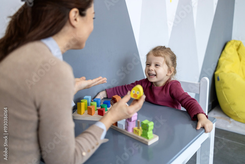 Preschool teacher engaging with a smiling toddler girl at a small desk, using wooden sorting blocks and a duck toy for cognitive development and speech therapy in a modern classroom