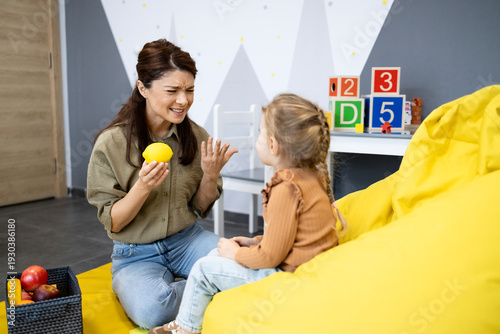 Speech therapist holding a lemon and making a sour face to a young girl, teaching her about taste and facial expressions during childhood development therapy