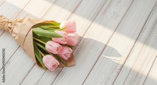 A small bouquet of pink tulips wrapped in brown craft paper rests on sunlit white wooden plank flooring.