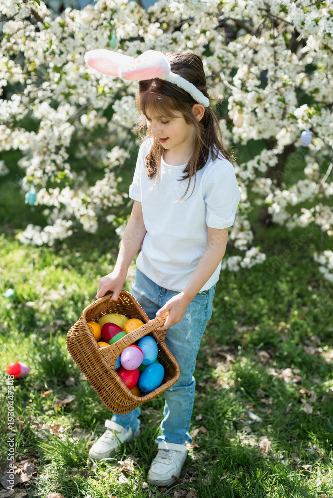 Fototapeta premium Easter egg hunt. Little girl with bunny ears holding decorative Easter egg and wicker basket under blooming spring tree. Outdoor Easter celebration. joyful childhood moment outdoors.