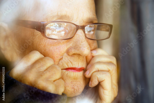Portrait of an old woman with glasses reflected in a window. The face of an elderly woman with glasses looking out the window.