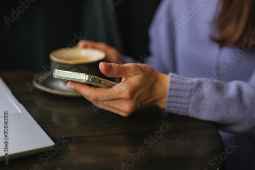 Close up of a woman in a purple sweater using a smartphone while sitting at a wooden table in a cafe with a moody, dark-toned interior.