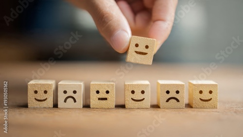  A wooden table surface. A row of wooden blocks showing feedback icons (sad, neutral, happy face). A hand is picking the happy face block. Customer satisfaction concept.