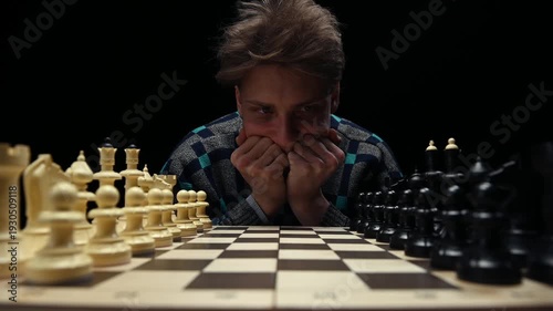 A young man is sitting in front of a chessboard with pieces and thinking about the next move. The chess player is assessing the risks before making the first move. A thoughtful chess player
