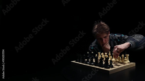 A young man is sitting in front of a chessboard with pieces and thinking about the next move. The chess player is assessing the risks before making the first move. A thoughtful chess player