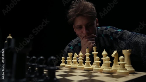 A young man is sitting in front of a chessboard with pieces and thinking about the next move. The chess player is assessing the risks before making the first move. A thoughtful chess player