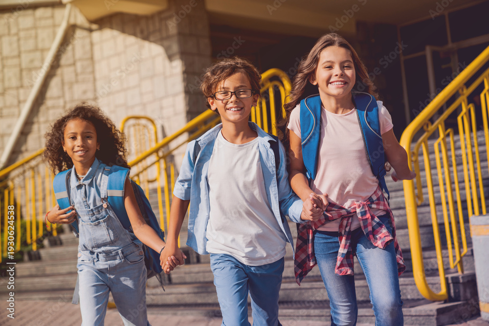 Obraz premium Happy kids elementary school pupils children with rucksacks backpack walking holding hands at school yard running out outside at the end of the lessons. Finish school and summer vacation concept.