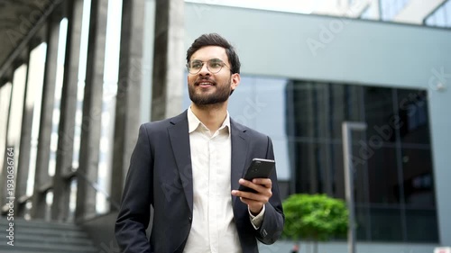 Confident businessman in a formal suit uses mobile phone while walking on the street near modern office building. Handsome male worker in glasses chats online or writes reads messages on a smartphone