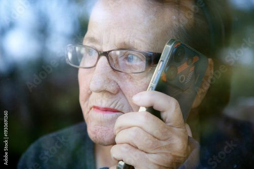 Portrait of an elderly woman with a phone in her hand at the window. An old woman with glasses is talking on her cell phone while looking out the window.