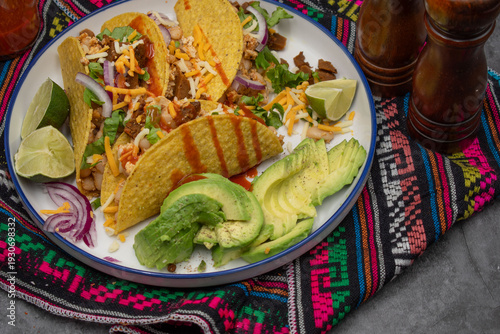 A vibrant vegan taco platter featuring soft tacos filled with seasoned seitan, tofu, and beans, served with fresh avocado slices, lime wedges, and sliced purple onion.
