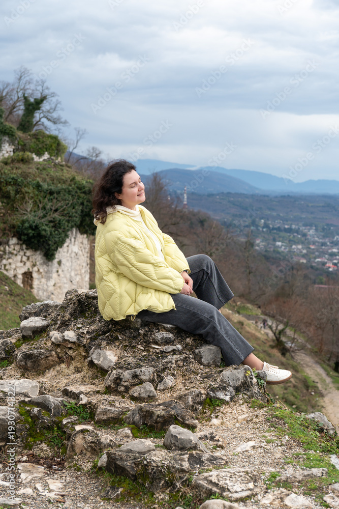 Fototapeta premium Woman sitting on stone ruins overlooking New Athos valley from Anacopia fortress in Abkhazia under cloudy sky, Travel lifestyle scene suitable for tourism marketing, outdoor destination promotion