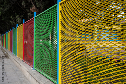 A vibrant, multi-colored fence made of expanded metal mesh panels in red, yellow, and green extends along a paved sidewalk in a linear perspective.