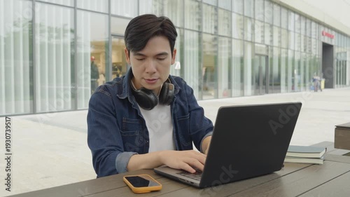 Young man in a denim jacket working on a laptop computer with a modern building in the background 