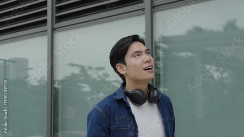 Young man with headphones in casual attire standing and looking upwards with expression of wonder in the urban environment