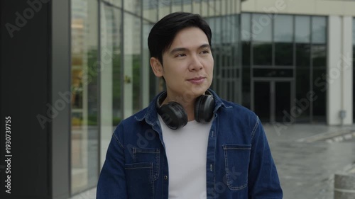 Young person with headphones is outside in front of the building. The person has a jacket and shirt 